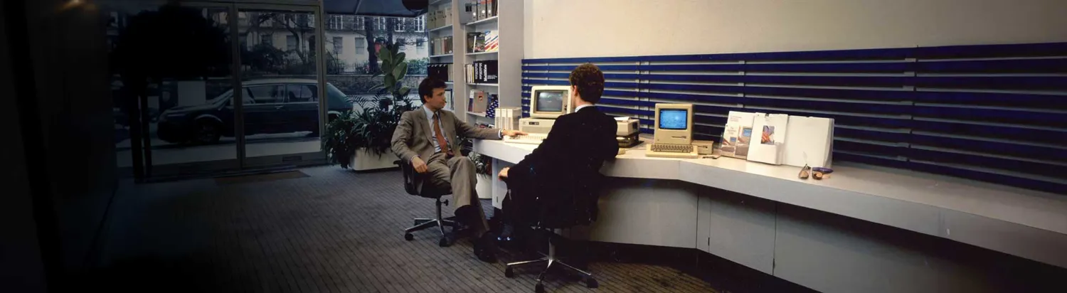 Two professionals engaged in a discussion at a modern office desk setup featuring vintage computer equipment