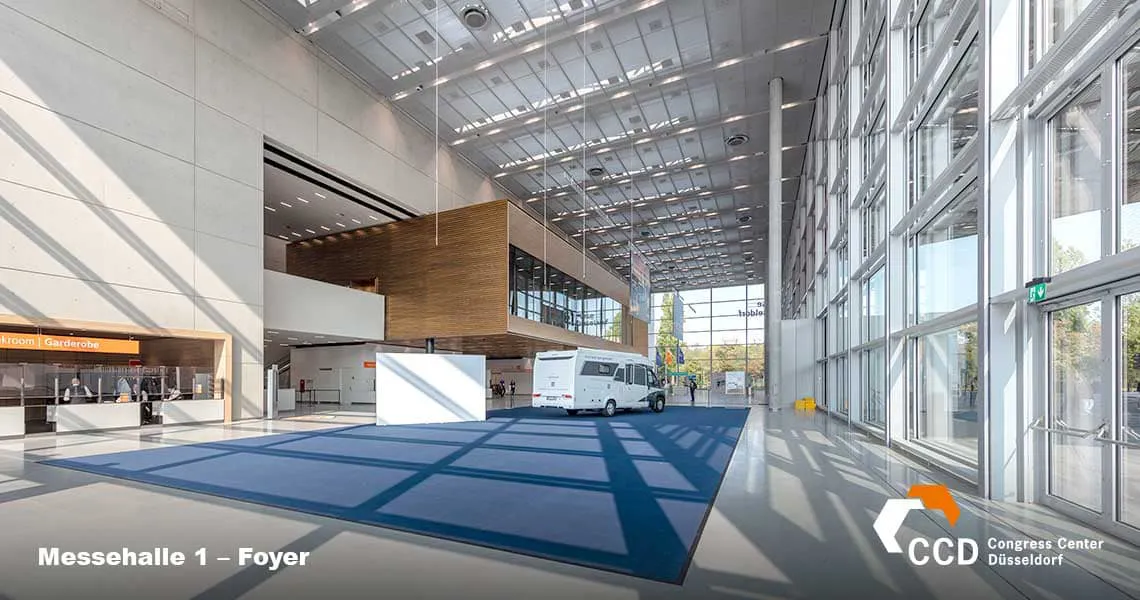 Large modern foyer of Messehalle 1 at the Congress Center Düsseldorf, featuring high ceilings, large windows allowing natural light, and a spacious blue-patterned carpet. Visible are a van parked near the entrance and a signpost directing to restrooms.