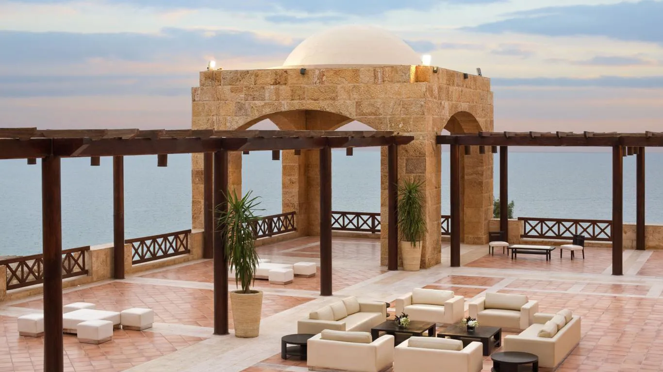 Elegant outdoor seating area with stone pavilion structure overlooking the ocean, featuring modern lounge furniture, wooden pergola, and potted plants. The setting includes a scenic seaside backdrop during twilight.