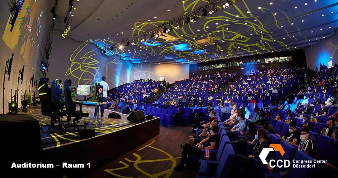 A spacious auditorium filled with attendees seated in rows, facing a stage with a presenter delivering a session. The backdrop features dynamic lighting and abstract designs, creating an engaging atmosphere.