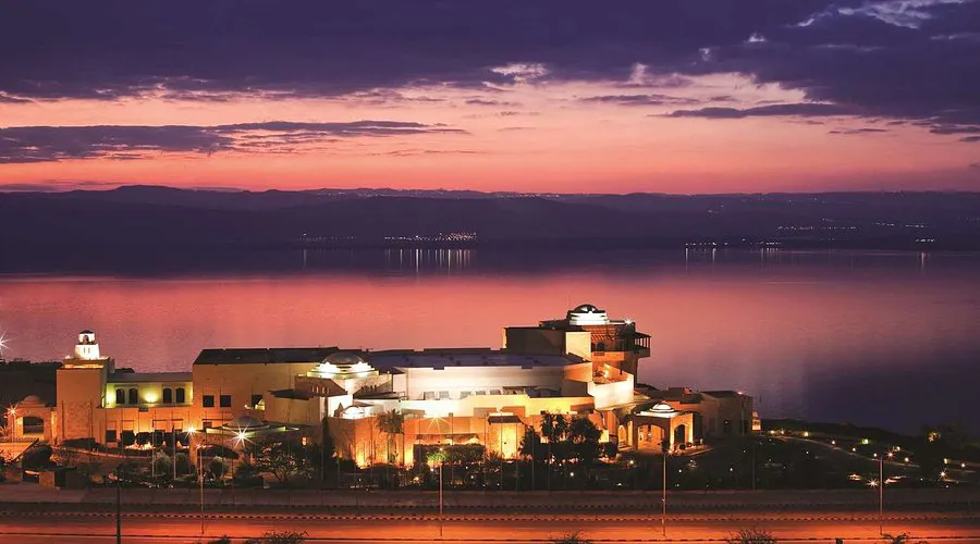 Elegant building complex illuminated by warm lights along a tranquil waterfront during twilight, framed by a vibrant sunset sky. The scene captures a serene, picturesque location ideal for grand events.