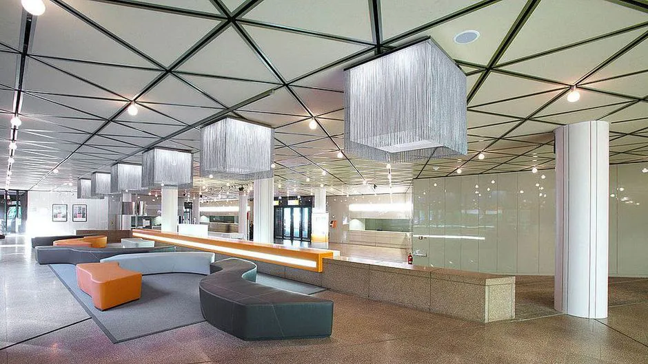 Elegant indoor conference or event space featuring modern seating arrangements with sleek, geometric pendant lighting suspended from an industrial ceiling. The area includes a reception desk with transparent partitions and a polished floor.
