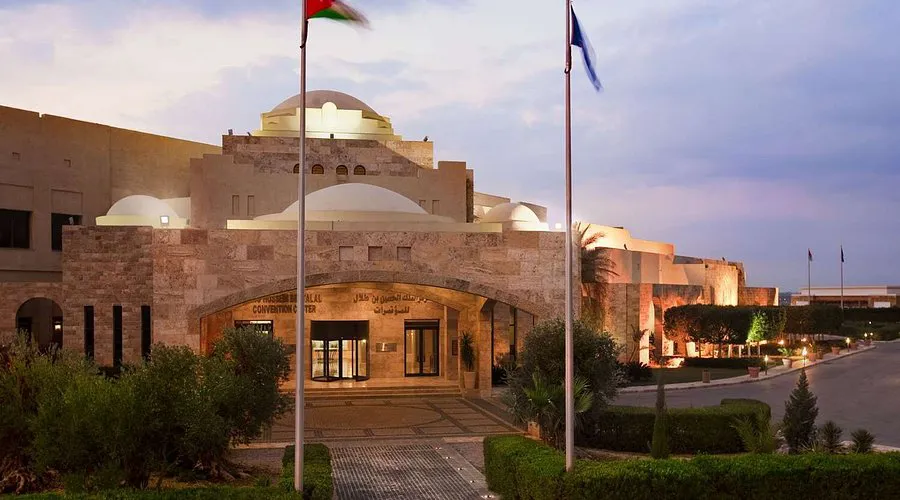 Elegant stone convention center building with modern architectural features, including domed structures and arched entryways, flanked by national flags. The scene captures the venue during twilight with well-maintained landscaping and illuminated pathways.