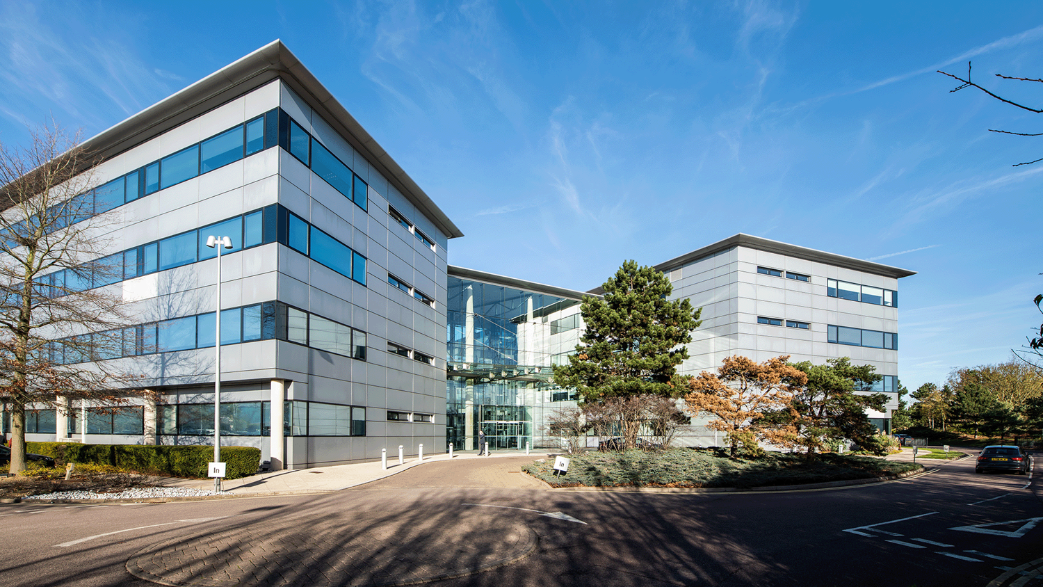 Modern office building with glass facades and sleek architectural design, featuring multiple floors and large windows under a clear blue sky. The entrance is flanked by landscaped greenery and a well-maintained pathway leading to the building.