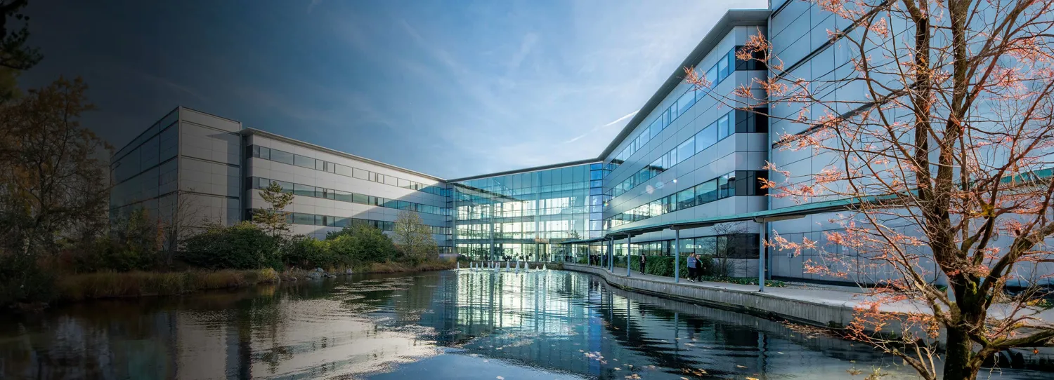 Modern office building with expansive glass facades reflecting a tranquil pond and surrounding greenery, framed by blooming trees with pink blossoms under a partly cloudy sky.