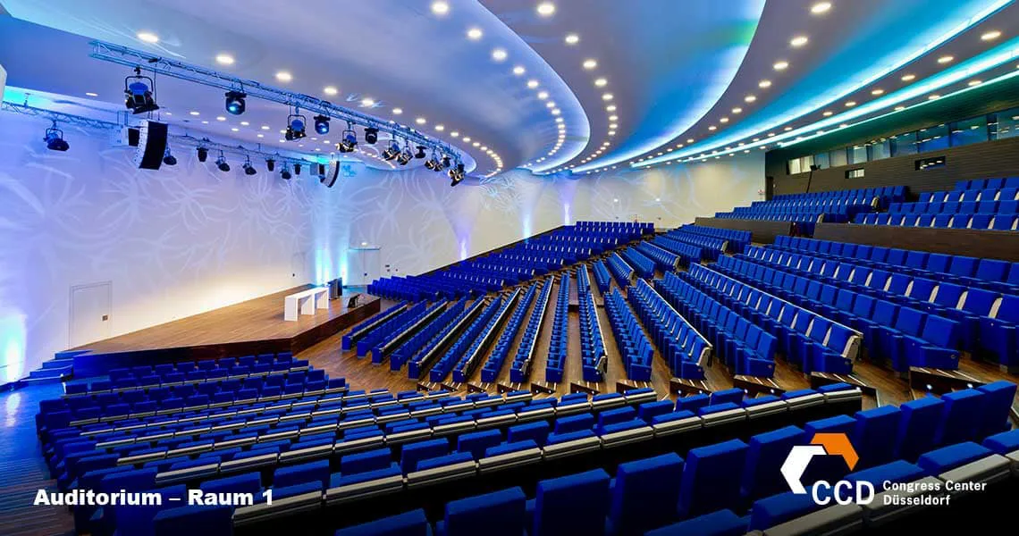 Large conference auditorium featuring blue seating arranged in rows facing a central stage area. The space is equipped with modern lighting and stage rigging, ideal for presentations and events.