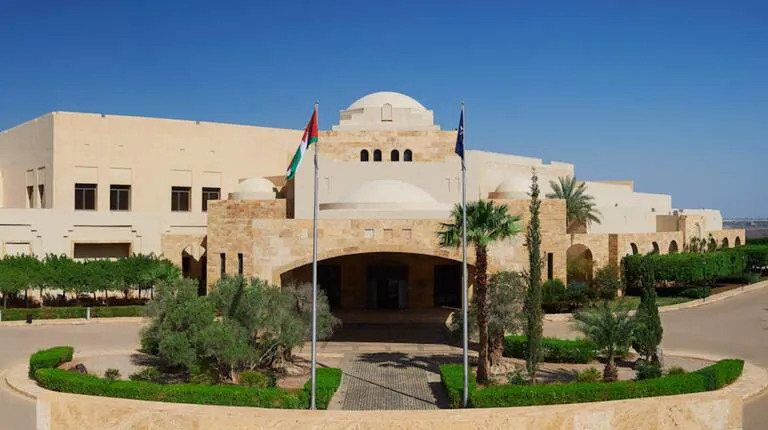 Elegant stone building with traditional architectural elements featuring domes and arched windows, surrounded by landscaped greenery and palm trees. Flags are prominently displayed at the entrance, indicating a formal and distinguished venue setting.