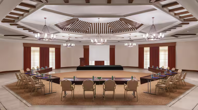 Elegant conference space featuring multiple round tables arranged in a circular layout, surrounded by modern chairs. The room includes a stage area with red drapes and stylish chandeliers hanging from a decorative ceiling.