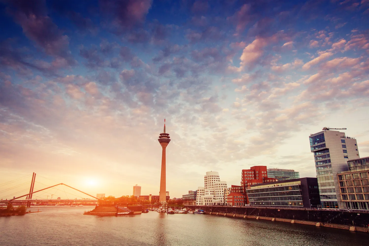Modern cityscape featuring a prominent observation tower along a riverfront during twilight, with contemporary buildings and a distinctive arched bridge spanning the water. The sky displays soft pastel hues blending with scattered clouds.