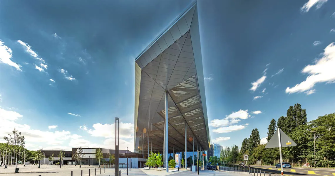 Modern architectural building with reflective glass facade rising against a bright blue sky. The structure features geometric design elements and is surrounded by landscaped greenery and urban infrastructure.
