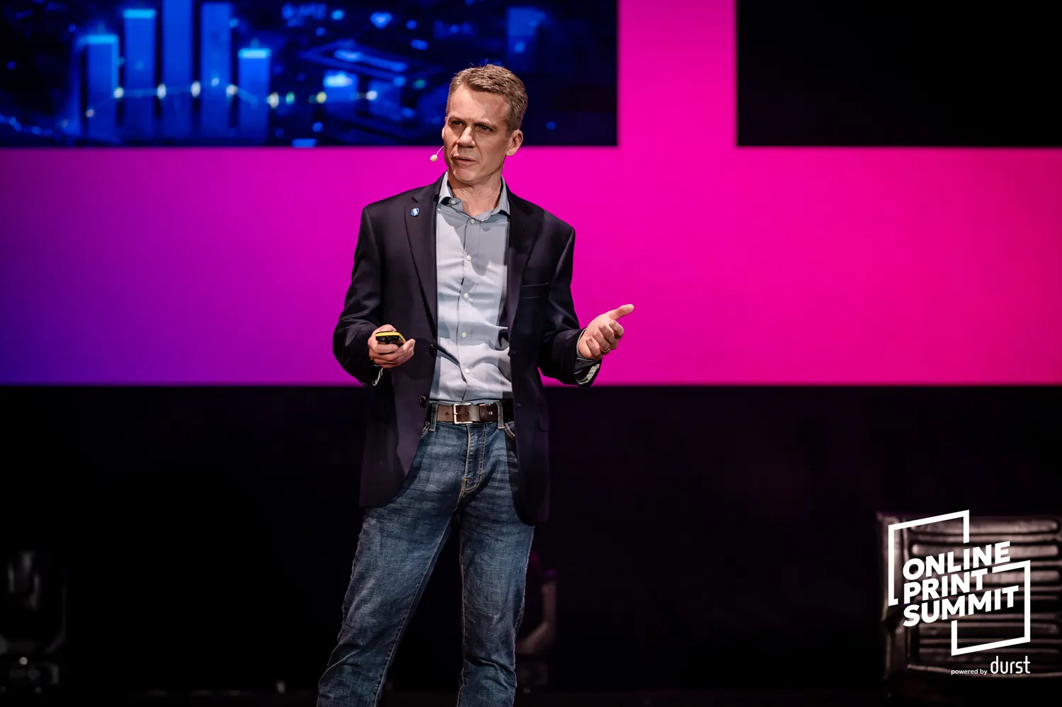 A man in a suit and casual trousers presents or speaks at a vibrant stage setting during an online event. The backdrop features dynamic lighting and branding for the Online Print Summit.
