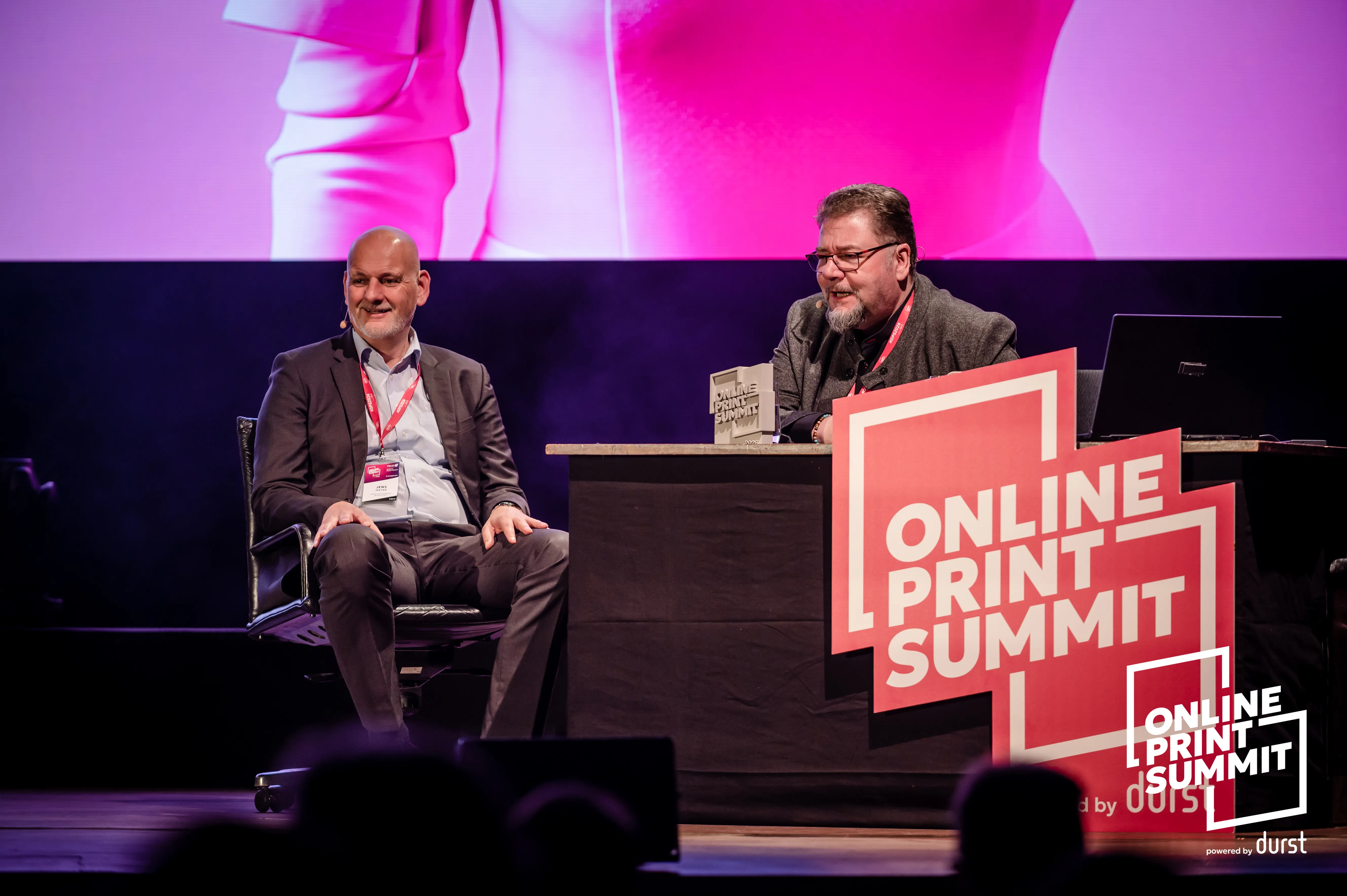 Professional panelists engaged in discussion at the Online Print Summit, featuring branded stage elements with bold typography and a backdrop of vibrant lighting. Attendees are seated at a table with laptops and promotional signage.