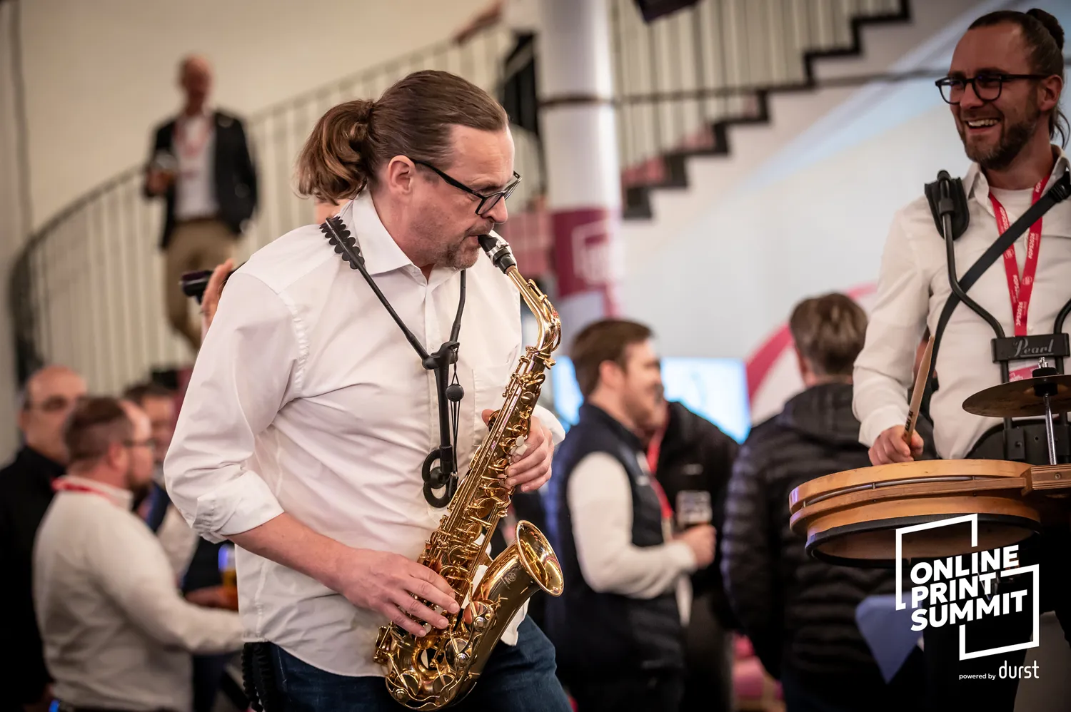 A saxophonist plays during an indoor event, surrounded by attendees. Another musician with drumsticks and headphones stands nearby, engaging in a lively musical performance.