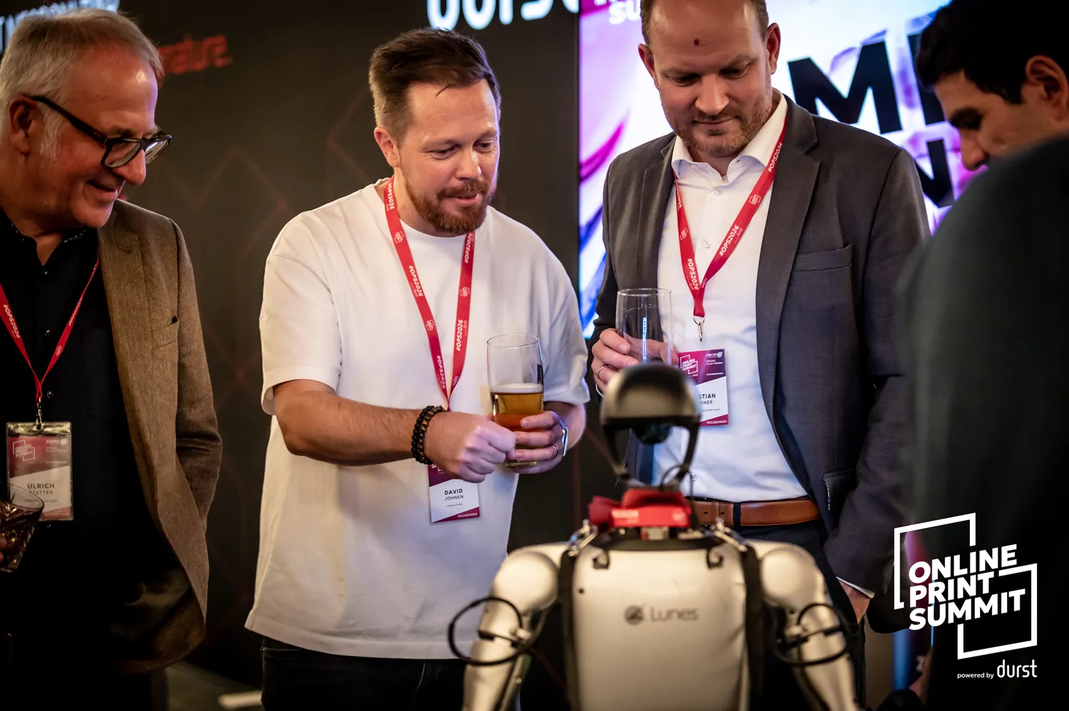 Professional attendees at a networking event holding drinks, engaged in conversation near a recording setup at the Online Print Summit. Lanyards and event badges indicate participation in the conference.