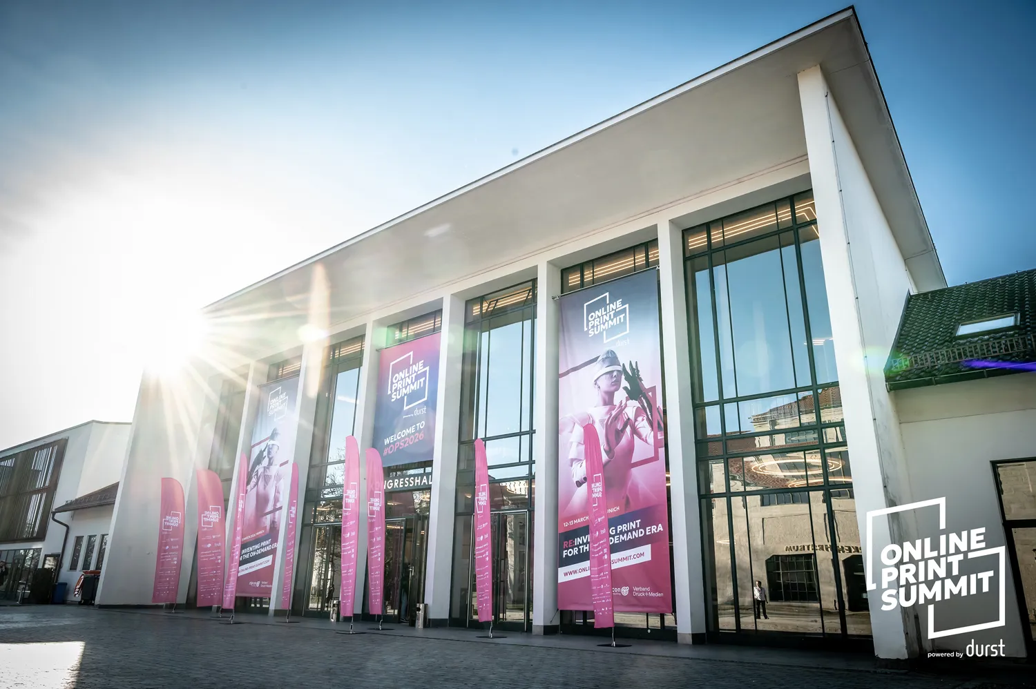 Modern conference building exterior with large glass entrance doors displaying promotional banners for the Online Print Summit. Pink vertical banners feature event branding, dates, and imagery of professional individuals. Sunlight illuminates the scene, highlighting architectural details and event signage.