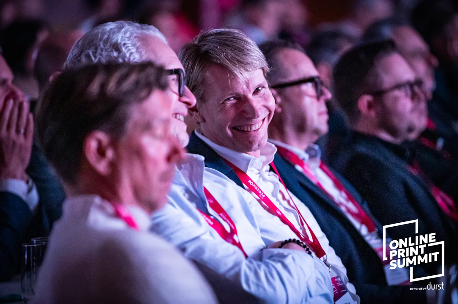 Professional attendees engaged in a lively discussion or presentation at an online industry summit, wearing lanyards with event branding. Warm lighting highlights their attentive and enthusiastic expressions.