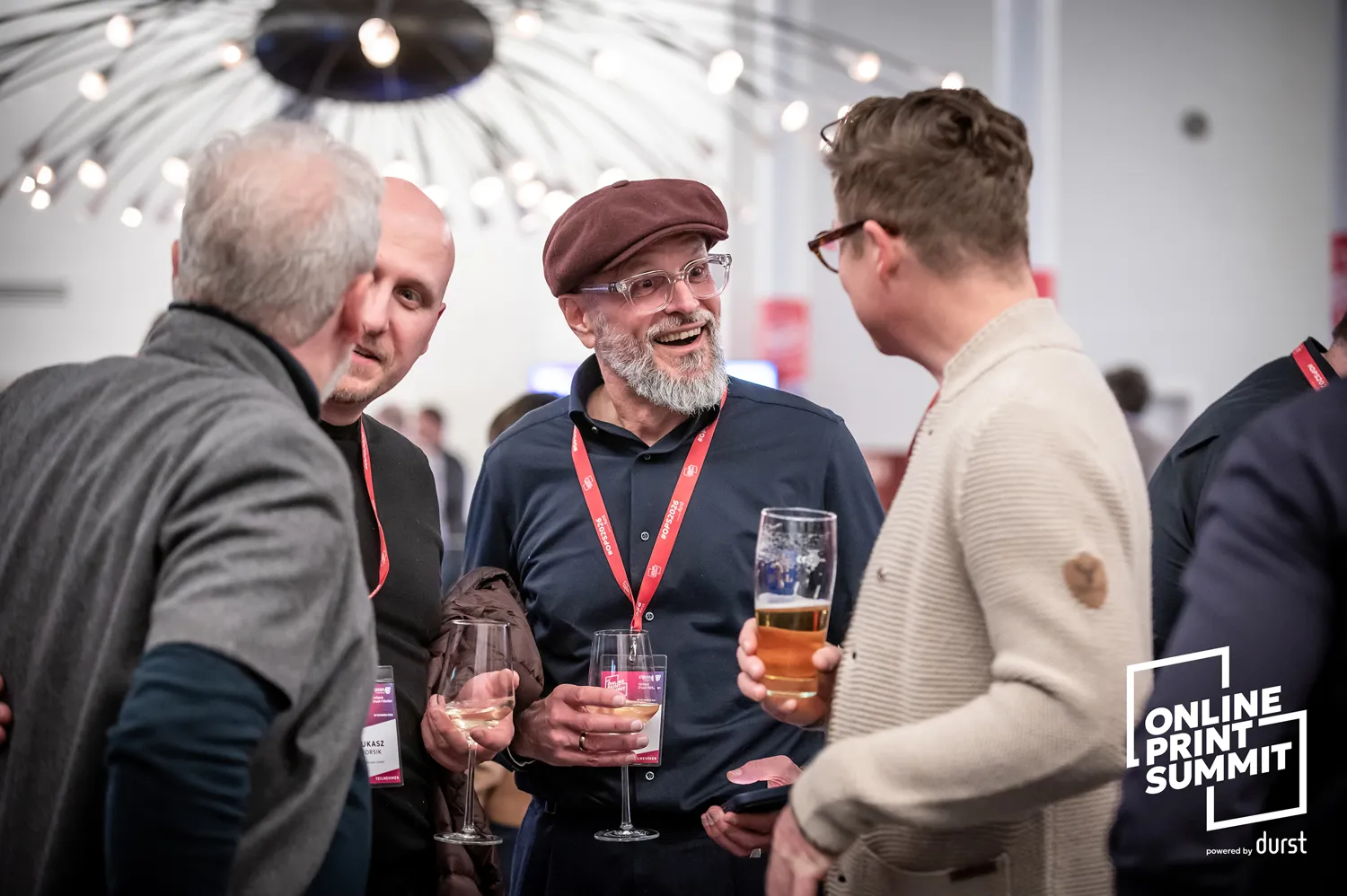 Attendees engaged in conversation at a professional networking event, holding drinks and wearing lanyards, set against an indoor venue backdrop with decorative lighting. Participants appear relaxed and engaged in discussions during a print industry summit.