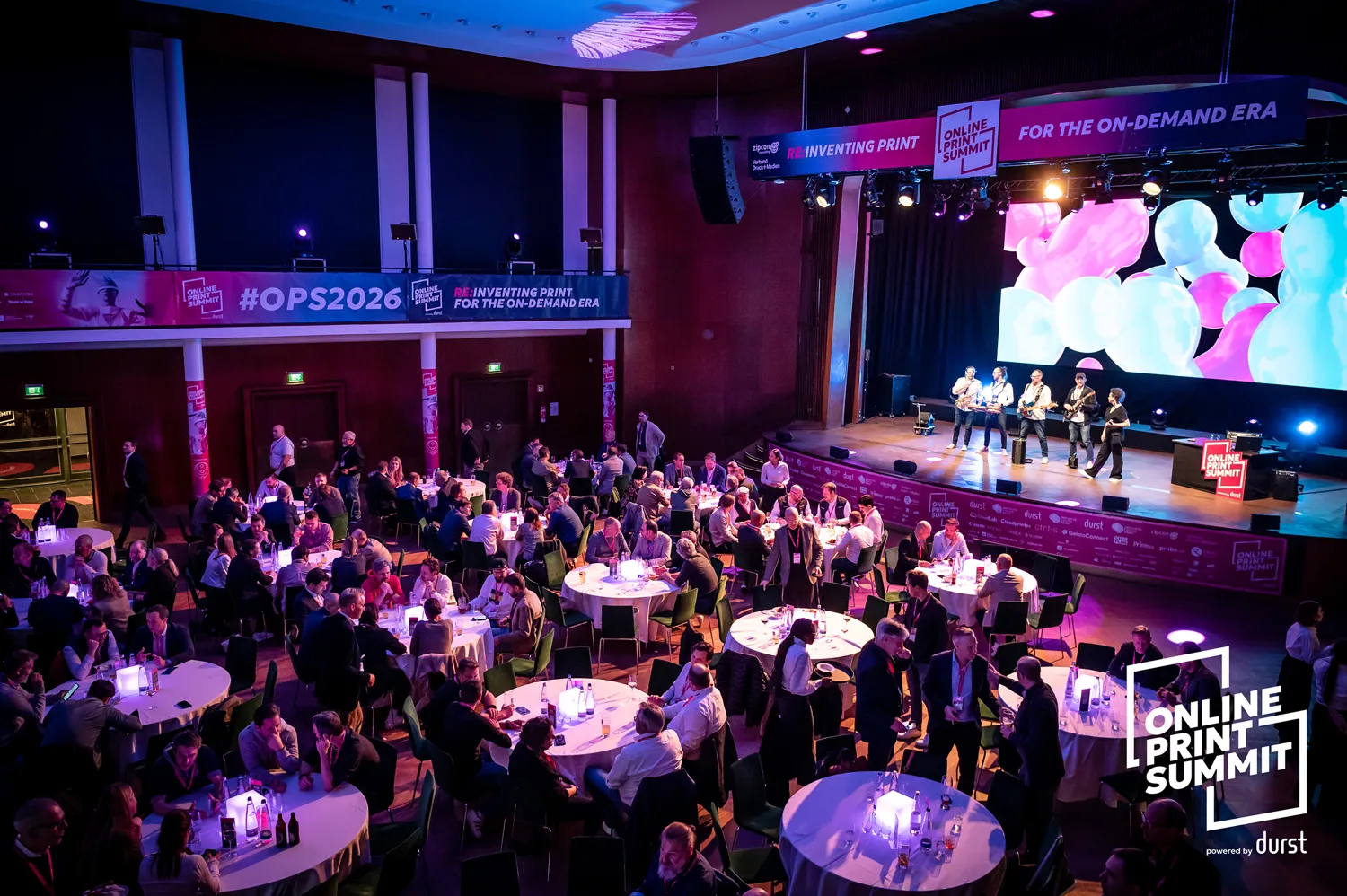 Professional attendees gather in a spacious event venue for an interactive session at the Online Print Summit. The scene includes round tables with participants engaged in discussions, a stage with presenters, and vibrant stage lighting displaying event branding.