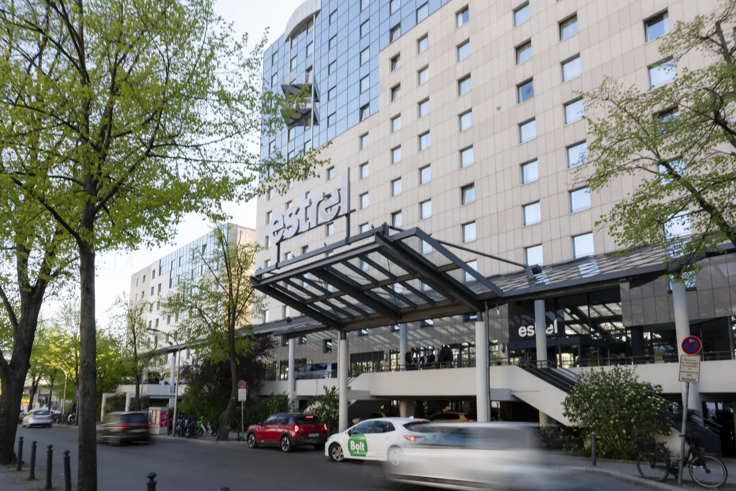 Modern multi-story hotel building with a covered pedestrian bridge entrance, featuring clean lines and large windows. The scene includes urban elements such as parked cars, bicycles, and a busy street with traffic.