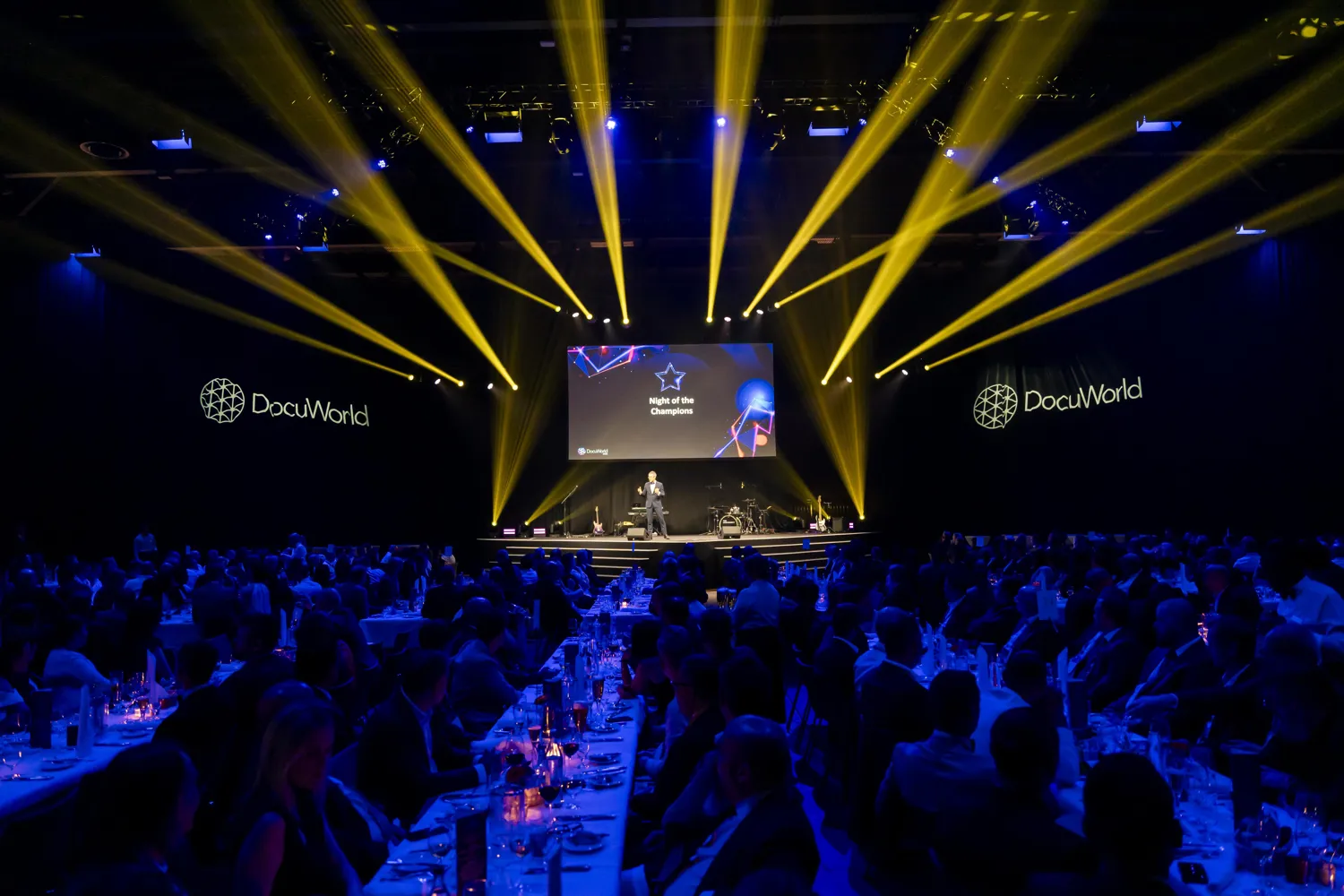 A vibrant gala event showcases an audience seated at tables under blue lighting, facing a stage. The stage displays the event branding "DocuWorld" and a screen announcing "Night of the Champions," with performers including a speaker and band members.