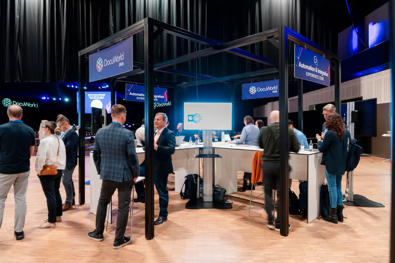 Professional attendees gather at a DocuWorld 2026 booth focused on automation and integration experiences, with interactive displays and presenters engaging with visitors in a well-lit exhibition hall. Booth signage prominently displays the event branding and session themes.