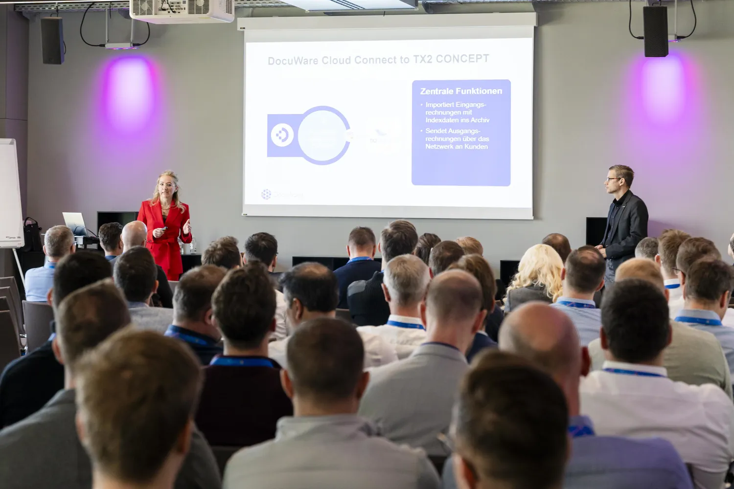 Professional seminar attendees engaged in a presentation discussing DocuWare Cloud integration with a focus on core functionalities, including data import and network-based output. A presenter in red speaks to the audience while another individual stands beside the screen displaying key concepts and visual workflows.