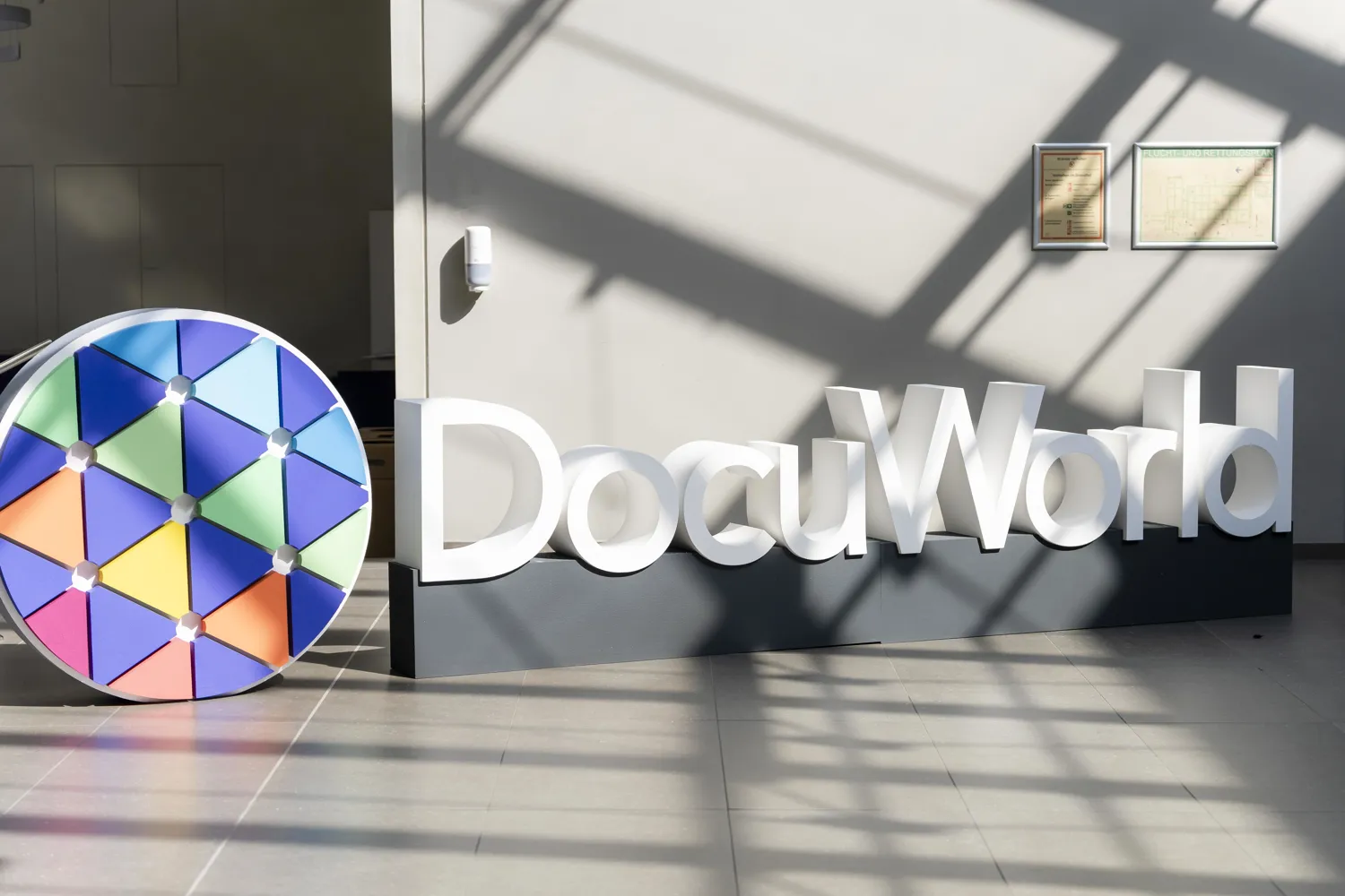 Multicolored geometric wheel sculpture positioned near a large, illuminated "DocuWorld" sign on an indoor entrance. The scene captures a modern, welcoming event venue with directional and branding elements.