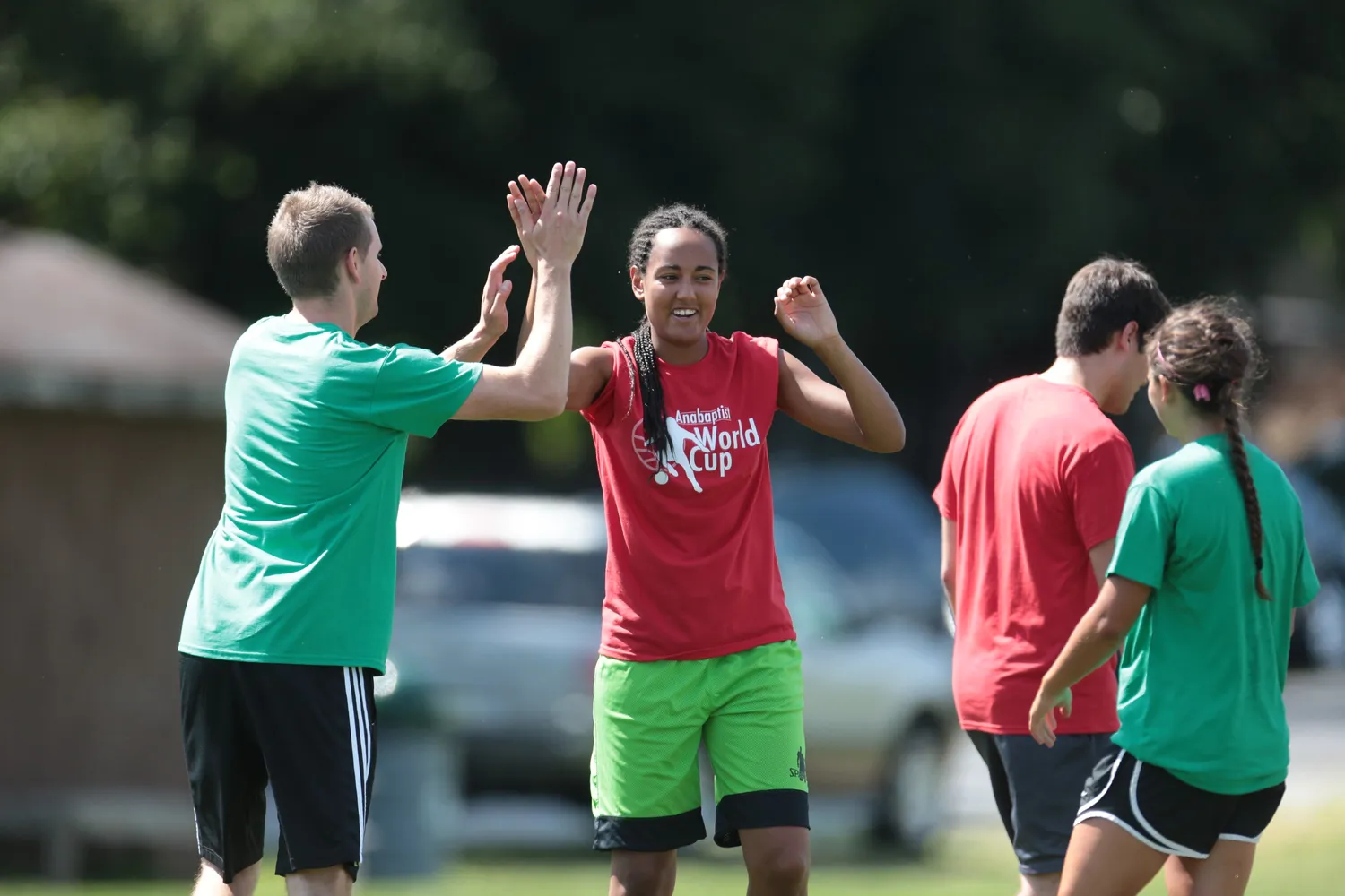 Athletes in athletic attire celebrating with a high-five on an outdoor sports field, wearing team jerseys and shorts, with one shirt displaying 'Anabaptist World Cup' branding.