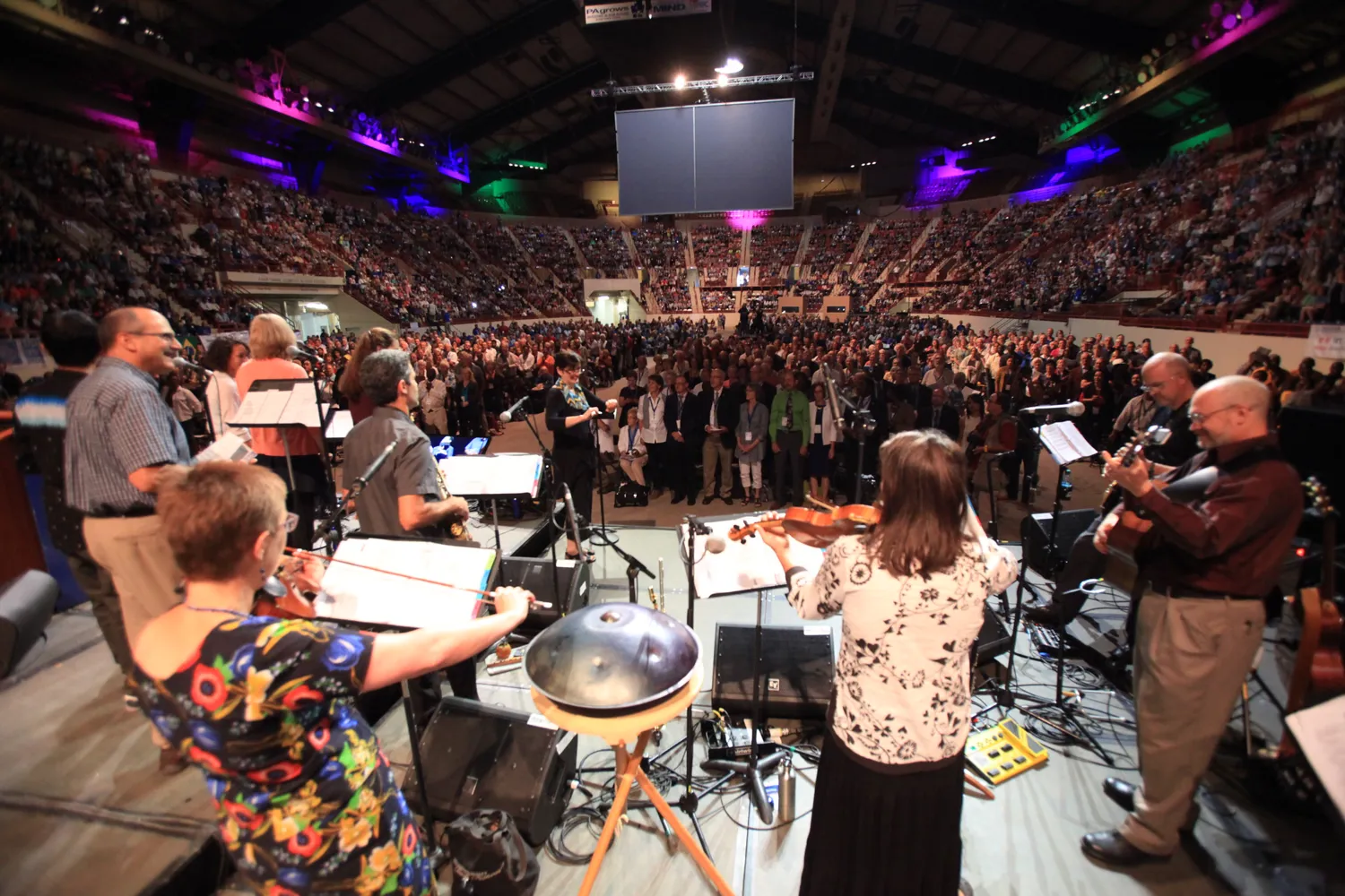 A sizable audience attends a live musical event in an indoor arena. Musicians perform on stage, including string instruments, percussion, and vocalists, with colorful stage lighting enhancing the atmosphere.