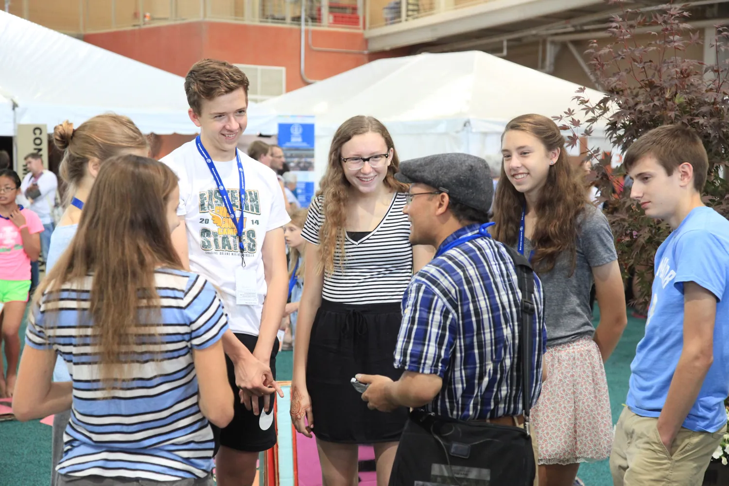 A lively outdoor academic conference scene features attendees engaged in conversation. Participants wear lanyards, casual attire, and some display event badges or shirts with text and logos. The group appears to be outdoors under tents with greenery in the background.