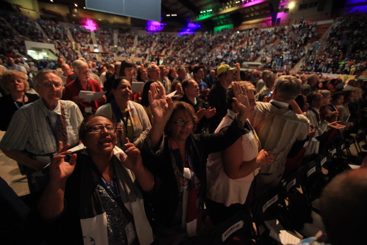 A large audience participates enthusiastically in a well-lit indoor event, engaging with a speaker or presentation. Attendees are seated closely together, holding papers and raising hands, indicating active involvement in the discussion or presentation.