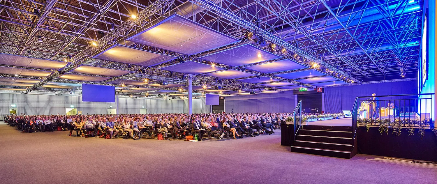 A spacious conference venue features an audience seated in rows facing a stage with professional lighting and a podium setup. The venue has an industrial metal truss ceiling with ambient stage lighting creating a formal and engaging atmosphere.