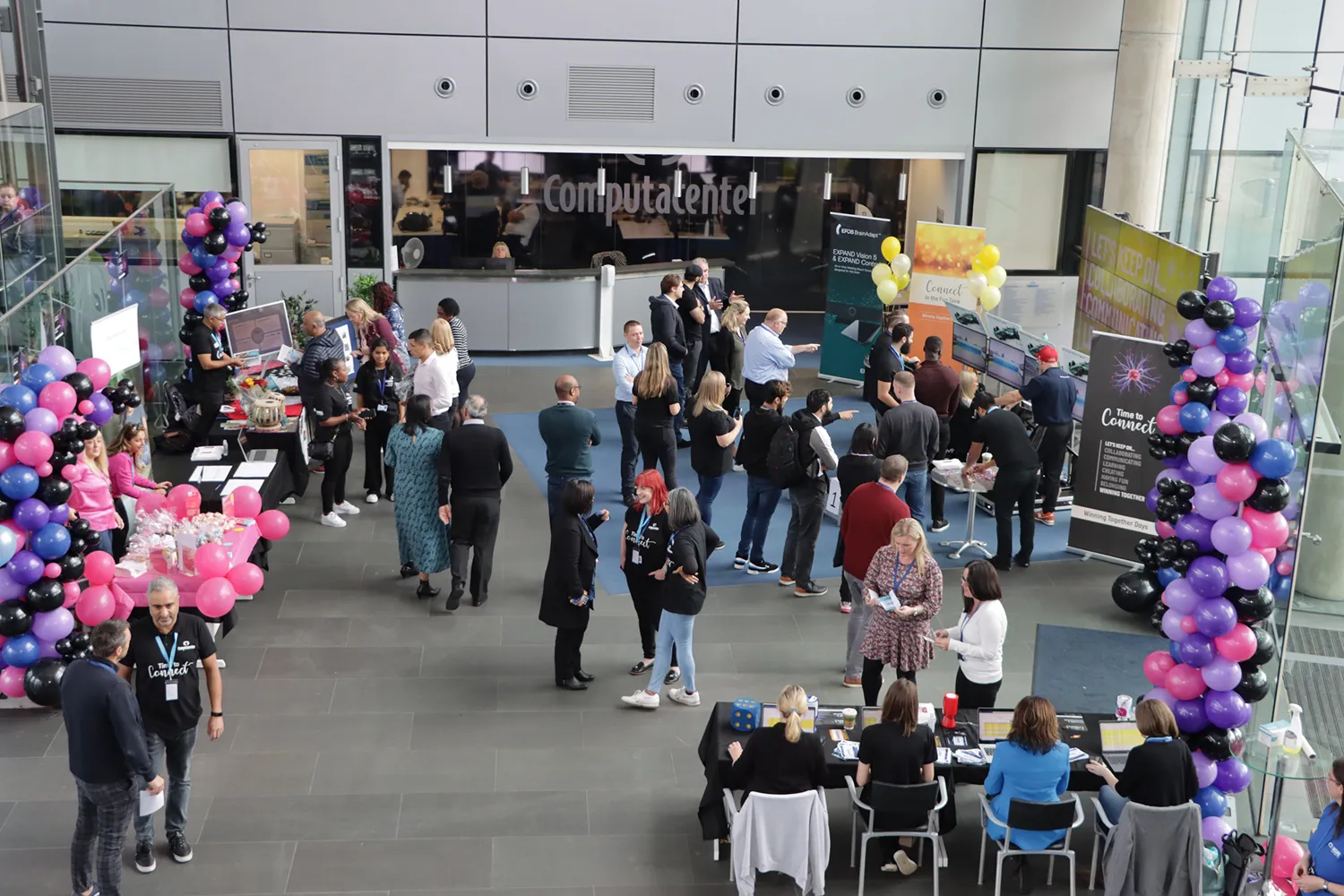 Attendees gather in a modern conference space for a professional networking event. Colorful balloons and branded booths highlight interactive stations with participants engaging in discussions and activities.