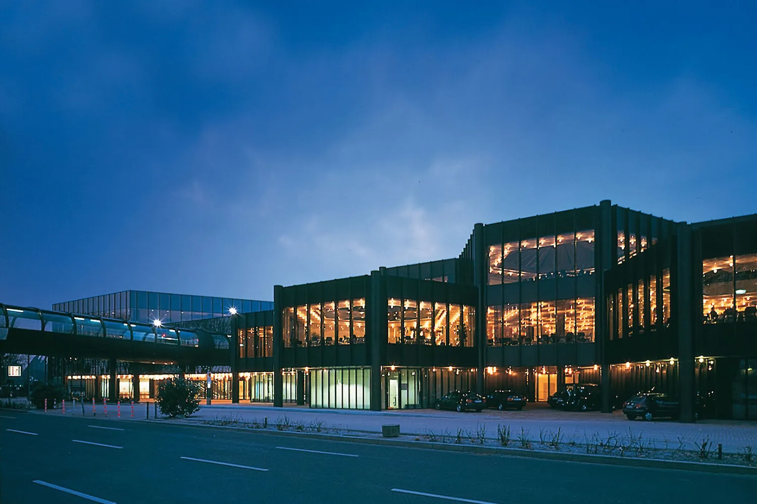 Modern convention center exterior illuminated at dusk with warm lighting reflecting off large glass windows, showcasing indoor event spaces. The scene captures a well-lit, professional venue with modern architectural design and an inviting atmosphere.