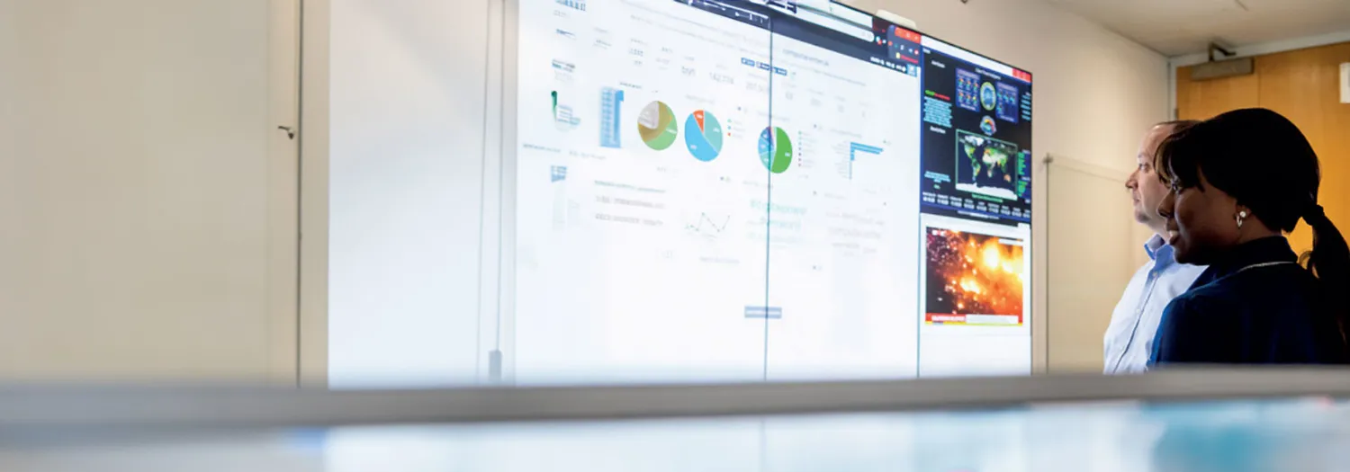 Two individuals are examining large digital displays filled with charts, graphs, and scientific visualizations in a conference or meeting room setting. The screens showcase statistical data and space-related imagery.