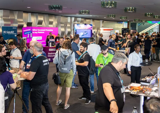 Professional attendees gather at a bustling tech expo, engaging with booths, discussing innovations, and exploring various exhibition displays. The scene captures a lively atmosphere with participants wearing event badges and casual business attire.