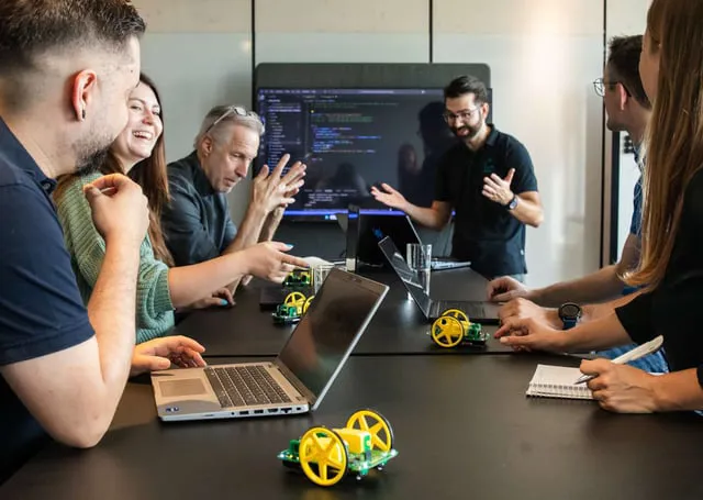 A group of professionals engages in a collaborative discussion around a table, surrounded by laptops and digital displays. They appear to be brainstorming or presenting technical concepts, with interactive elements like robotic models and notebooks present.