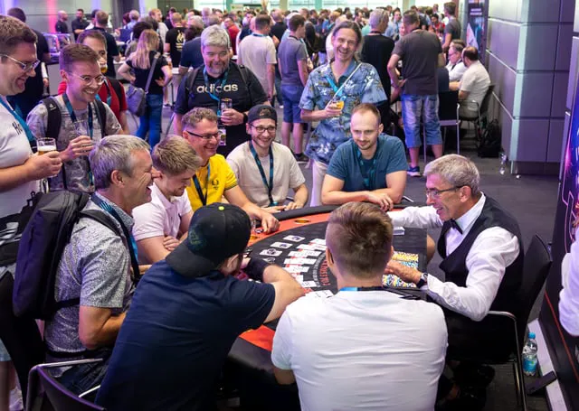 Attendees engaged in casual social interaction and tabletop gaming at a busy conference or trade show venue. A diverse group of individuals, some wearing lanyards and casual attire, participate in a card game while others socialize around them.