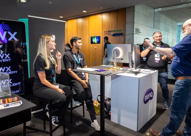 Attendees at a professional event engage with a tech demonstration booth featuring branded displays and interactive equipment. Three individuals interact with a Genio-branded setup, while another person stands nearby, gesturing and discussing the demonstration.