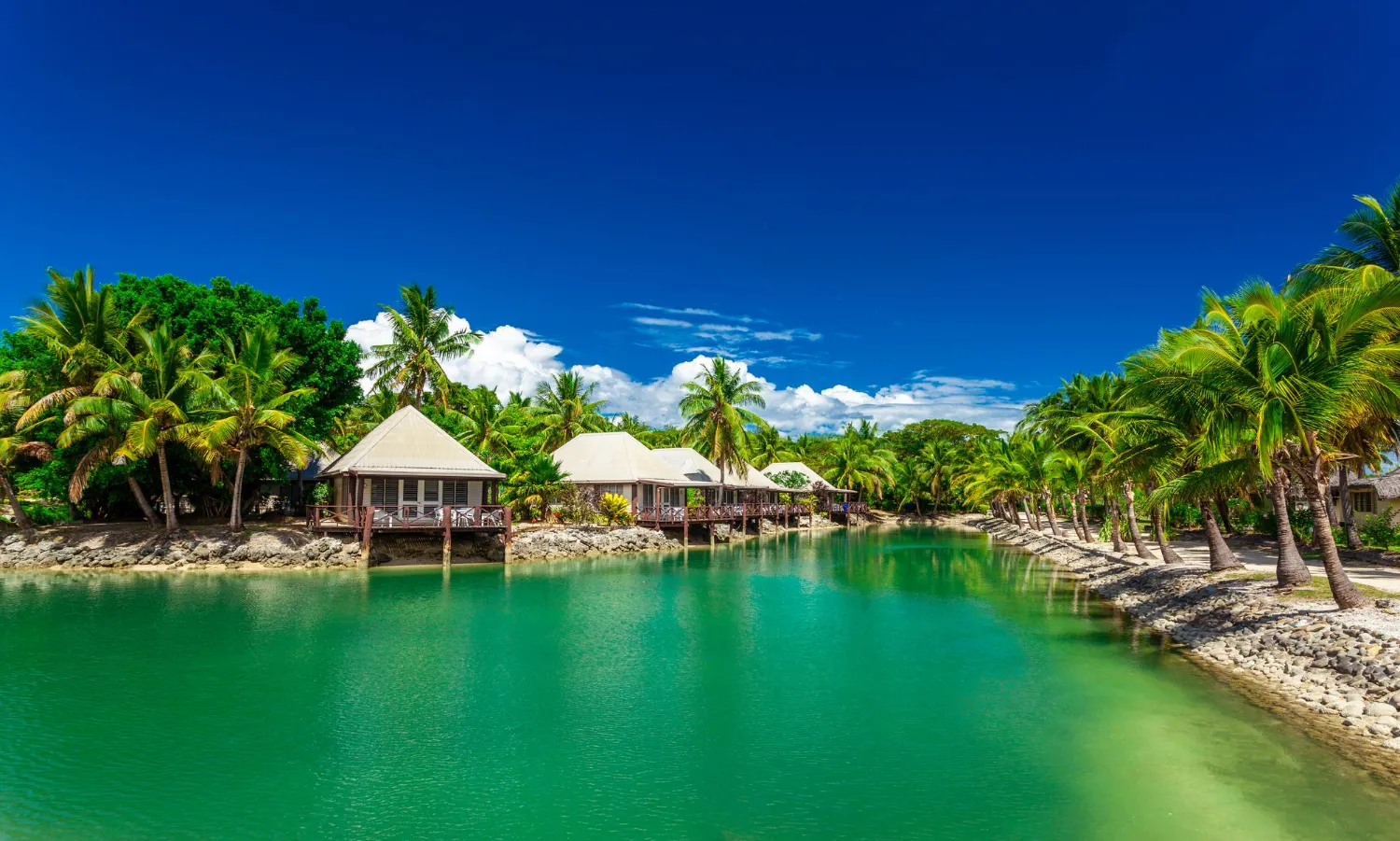 Lush tropical resort buildings with thatched roofs overlooking a calm, turquoise lagoon surrounded by dense palm trees under a bright blue sky.