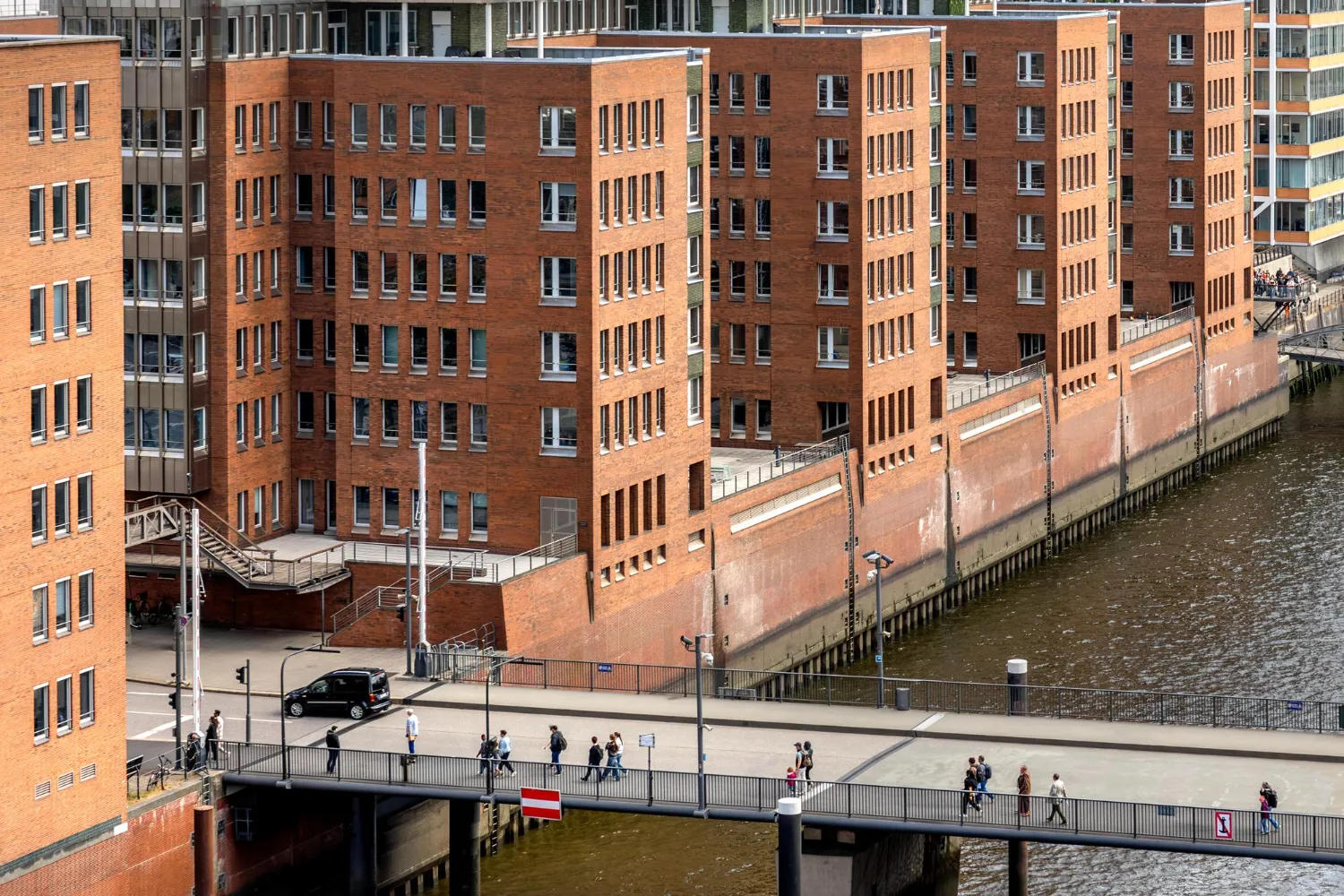 Multi-story brick buildings along a waterfront with multiple windows and balconies. A pedestrian pathway runs parallel to the water, with people walking and gathering near the bridge and riverbank.