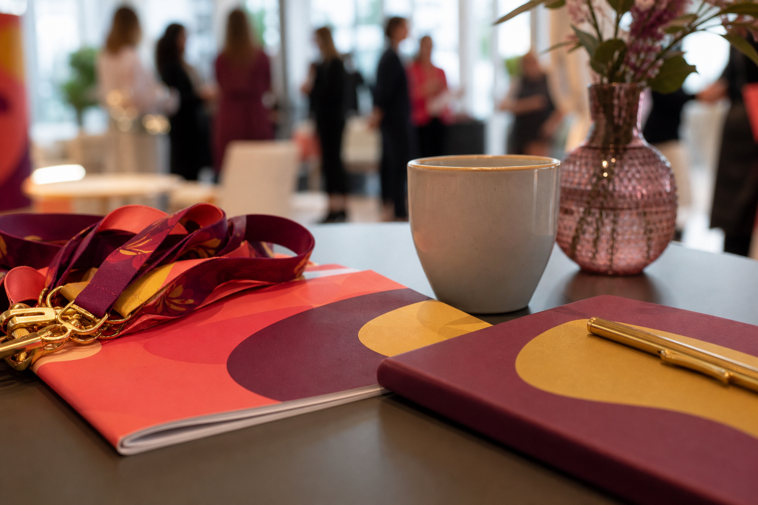 A table setup featuring branded notebooks with decorative ribbons, a ceramic mug, and a decorative glass vase with floral arrangement. Attendees in the background appear to be engaged in a networking or conference event.