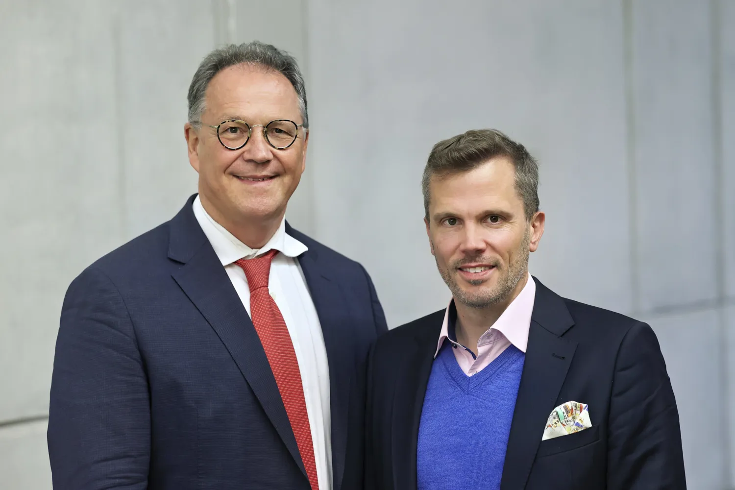 Two men in formal attire pose together outdoors against a neutral background. Both are wearing suits, ties, and pocket squares, exuding a professional and collaborative demeanor.