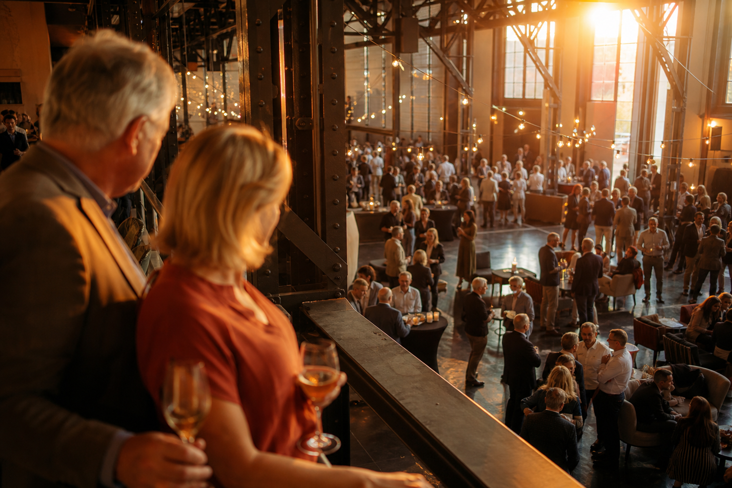 Attendees gather in a spacious, warmly lit venue with industrial architectural elements, engaging in conversations and enjoying beverages. Warm string lights and natural light from large windows enhance the cozy, social atmosphere during an organized event.