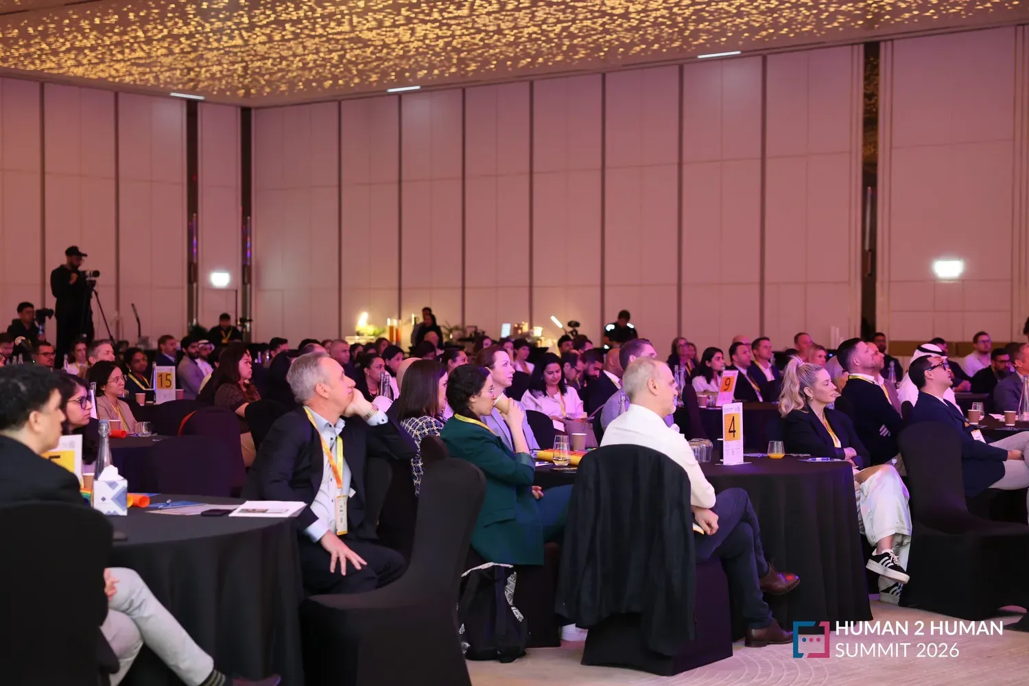 Rows of attendees at round tables facing the stage in a full ballroom at the Human2Human Summit 2026