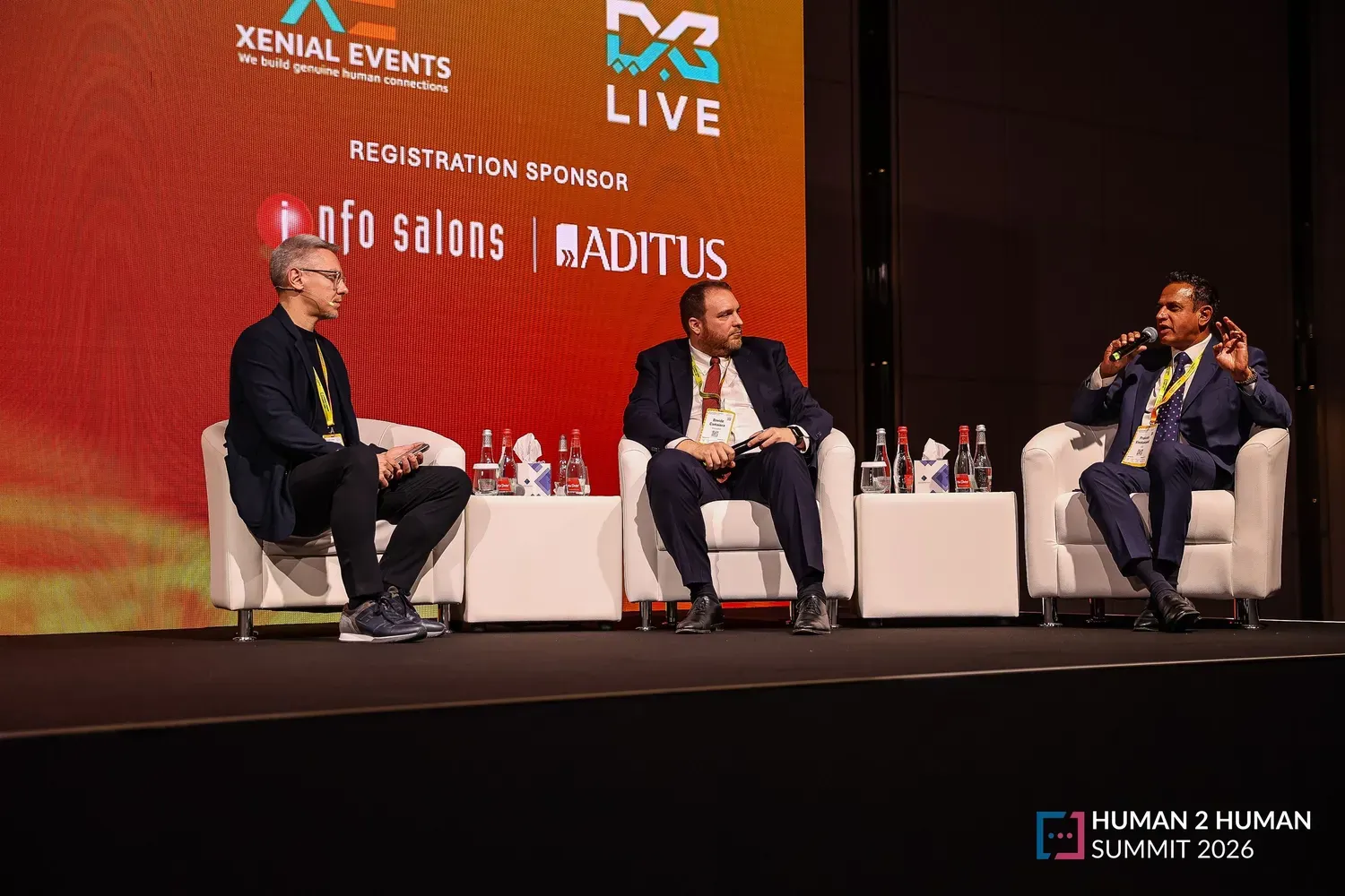 A three-person panel seated in white lounge chairs on stage, one panellist speaking into a microphone