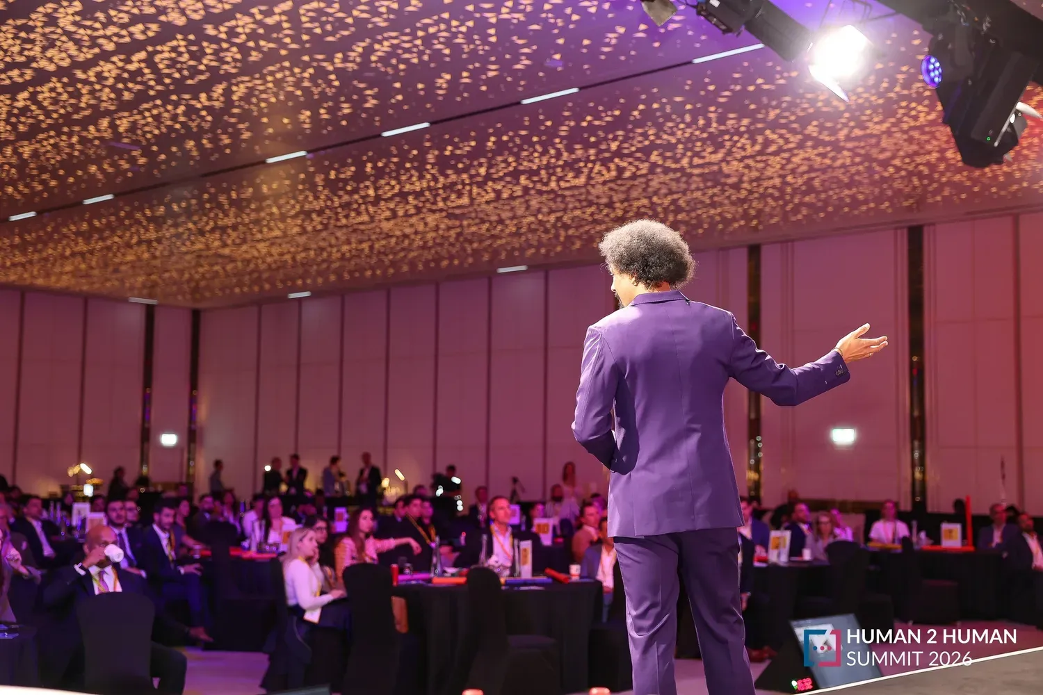 A presenter in a purple suit, seen from behind, addresses a full ballroom of attendees at round tables under a sparkling ceiling at the Human2Human Summit 2026