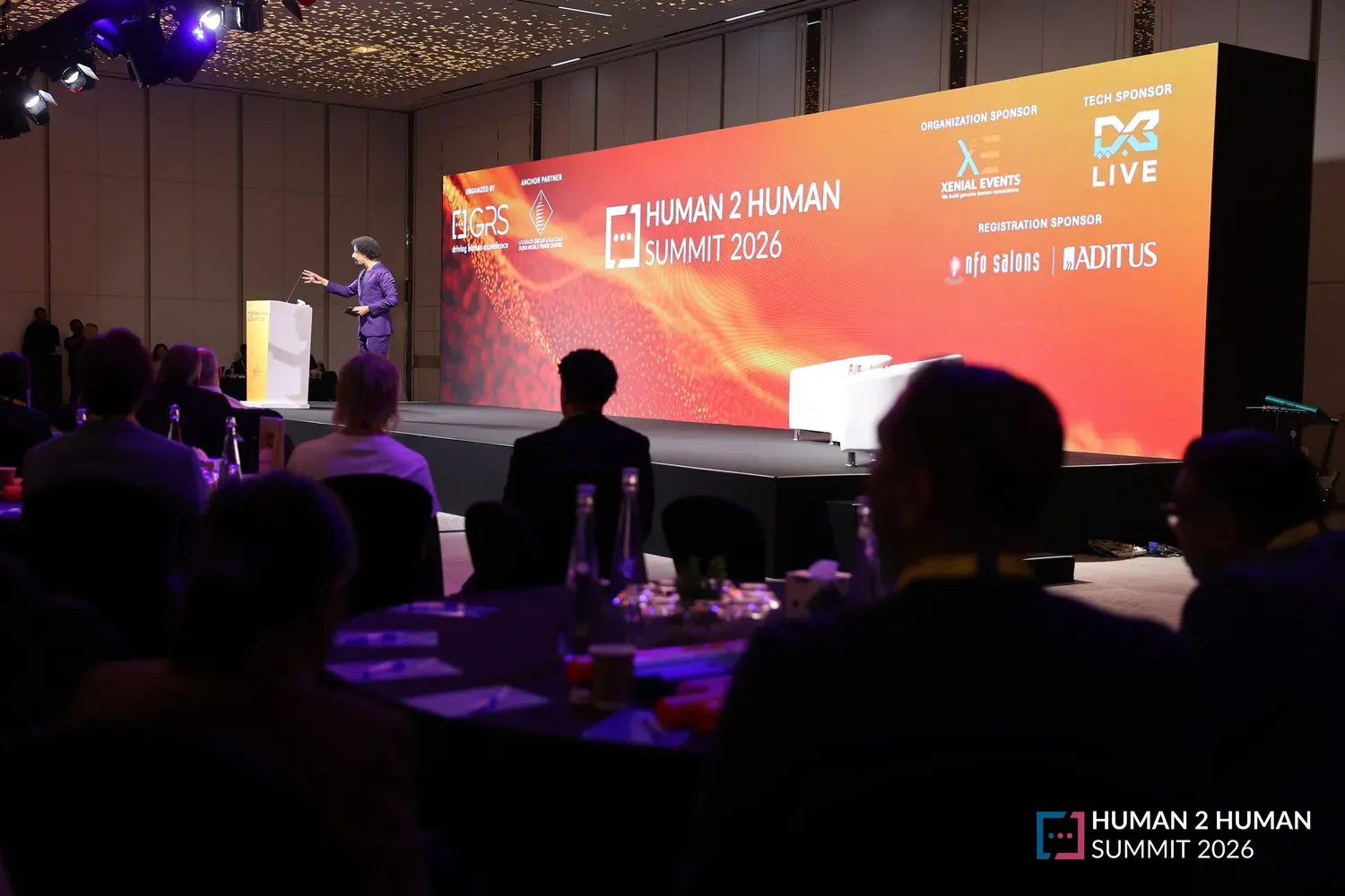 Wide view of the main stage at the 2026 summit: the host at a lectern before a huge red H2H screen, the audience seated at round tables in the darkened ballroom