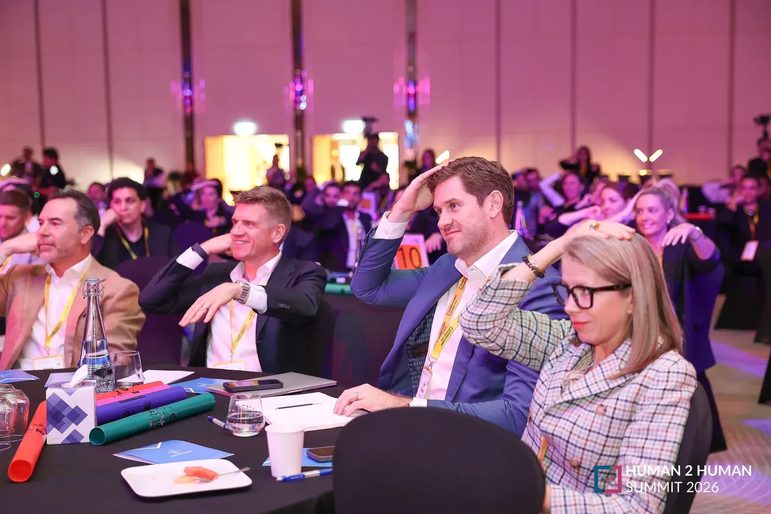 Smiling attendees seated at round tables with colourful props, engaged in a session at the 2026 Human2Human Summit