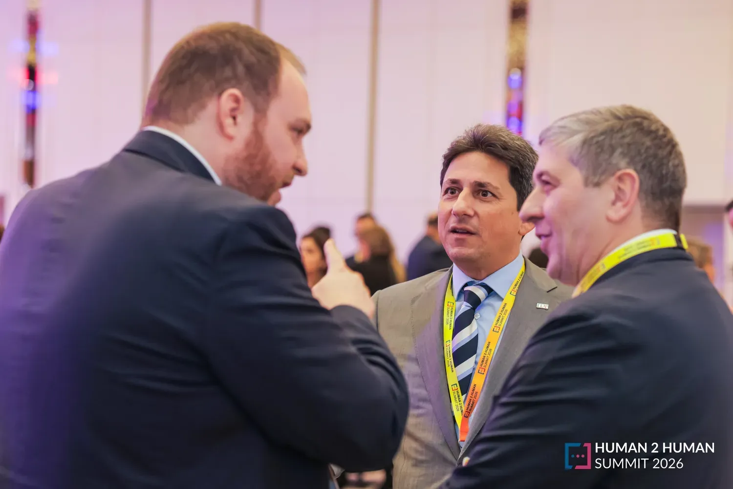 Three men in suits with event lanyards in animated conversation during a networking break